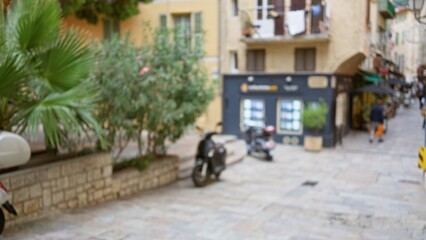 Blurred view of an outdoor street scene in villefranche-sur-mer, france, showcasing defocused architecture, foliage, and pavement in a sunny european town setting.