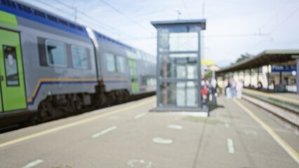 Obraz premium Blurred scene showing people waiting at a busy train station platform with a moving train in the background under bright daylight.