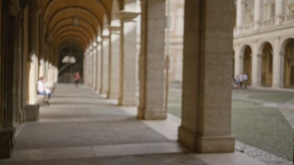 Obraz premium Blurred people walk through historic university campus with arches and stone columns creating a peaceful academic atmosphere outdoors under soft sunlight.