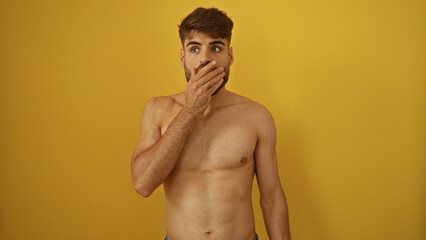 Fototapeta premium Young man with a beard looking surprised, covering his mouth, against an isolated yellow background