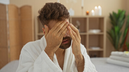 Young man with beard in spa room with hands on face wearing white robe during relaxation session