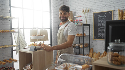 Handsome young hispanic man with beard smiling while working in a bakery, surrounded by fresh pastries and breads displayed in an indoor shop setting