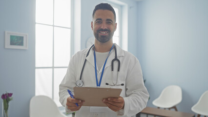 Handsome hispanic man doctor smiling in a hospital room holding clipboard