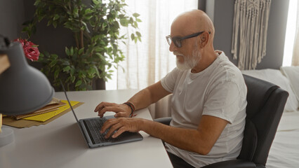Grey-haired man with a beard typing on a laptop in a bedroom with plants and decor, creating a cozy and productive home environment.