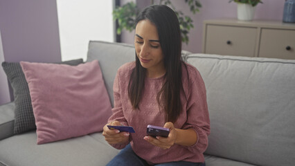 A young hispanic woman engages with a credit card and smartphone while comfortably seated on a living room couch indoors.