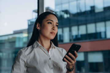 Asian businesswoman with smartphone standing by modern office window