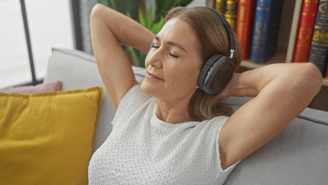 A mature woman relaxing with headphones on a sofa indoors, exuding tranquility and leisure in a comfortable home setting.