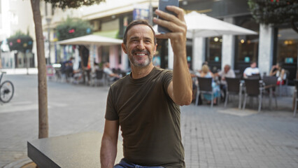 Hispanic man taking a selfie outdoors in an urban street setting, smiling happily with people dining in a background cafe.