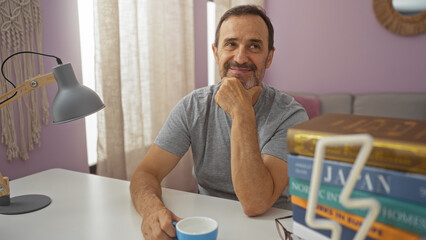 Handsome hispanic man indoors sitting in living room with a cup of coffee and books in front of him