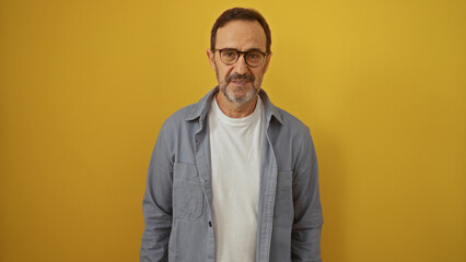 Fototapeta premium Middle-aged hispanic man standing isolated against a yellow wall, wearing glasses and a casual shirt, looking content.