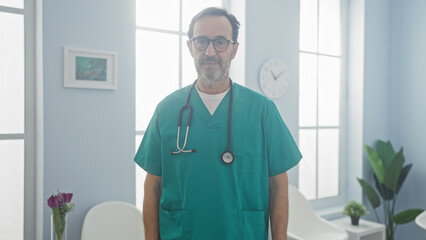 Mature hispanic male doctor in scrubs stands confidently in a bright hospital room.