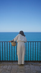 A young hispanic woman gazes at the expansive ocean, standing on a viewpoint in polignano a mare, puglia, italy, under a clear blue sky.