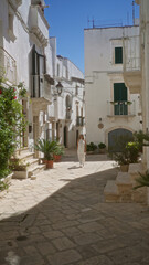 Naklejka premium A beautiful young hispanic woman walks through the charming old town streets of locorotondo in puglia, italy, surrounded by white buildings and lush greenery.