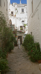 Young woman walking through the charming white streets of locorotondo, puglia, italy, adorned with vibrant plants and flowers in the old town on a sunny day.