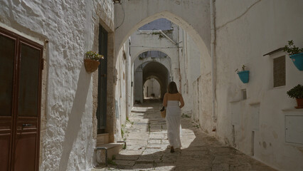 Naklejka premium Young hispanic woman walks through the charming narrow streets of ostuni's old town in puglia, italy, enjoying the historic whitewashed architecture and quaint atmosphere.