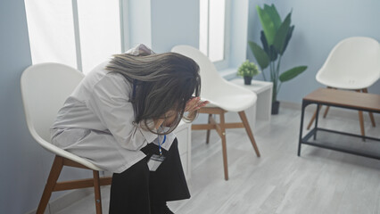 A young woman nurse sits in a clinic waiting room stressed and overwhelmed, covering her face in a moment of despair, surrounded by empty chairs in a bright, modern indoor setting.