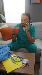 Obraz premium Young man with braids talking on phone and holding mug while wearing medical scrubs in cozy living room with backpack and stethoscope on table.