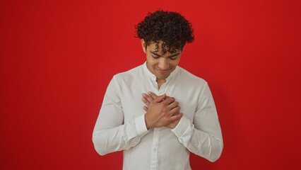 Young hispanic man with hands on heart, smiling, isolated on red background.