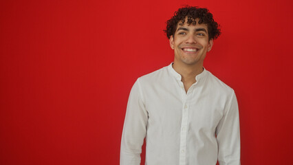 Fototapeta premium Smiling young hispanic man in white shirt standing against an isolated red background