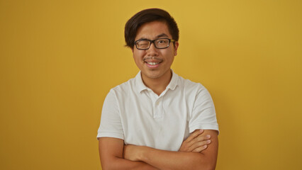 Obraz premium Young, handsome, chinese man in a white shirt smiling and winking while standing with folded arms against a vibrant yellow background