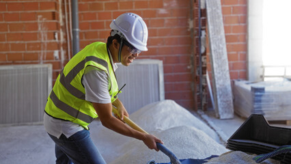 Young, chinese, man working indoors at a construction site, shoveling materials with a smile, wearing a hard hat and safety vest.