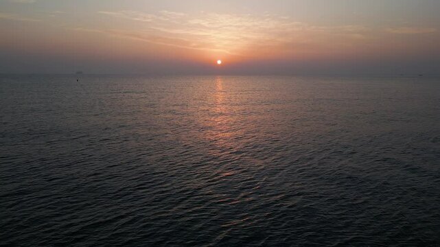 Aerial drone shot of a tranquil sea at sunset with gentle waves and a warm sun descending on the horizon, creating a soothing and picturesque ocean view ideal for relaxation themes