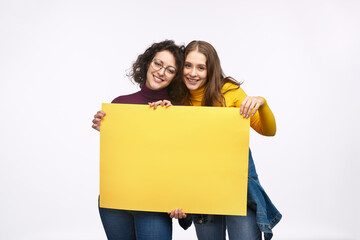 Two cheerful friends holding a blank yellow poster, showing unity and friendship. Ideal for...
