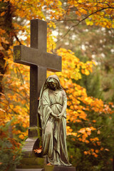 a female cemetery statue with her face black from weathering leans with folded hands against a cross in front of glowing autumn leaves in the background