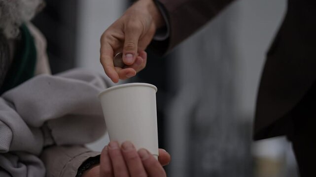 Contrast between wealth and poverty as businessman donating coins to homeless beggar cup in moment of giving and receiving. Closeup hands of business man dropping coins into homeless beggar cup.