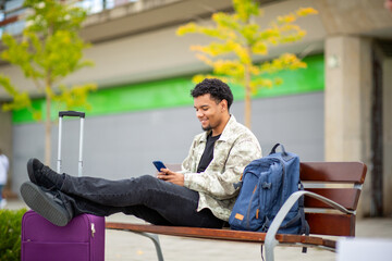 Man sitting on bench with smartphone, suitcase, and backpack