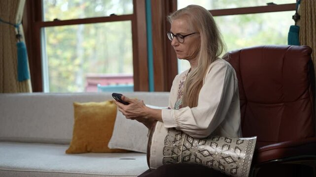 A woman with glasses and long blonde hair sits in a brown leather chair, holding a darbuka drum decorated with intricate patterns and playing it with her hand. There is a white couch and a window behi