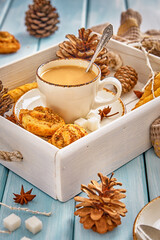 Coffee with milk and cookies, decorated with pine cones and branches on a white wooden tray on a blue table.