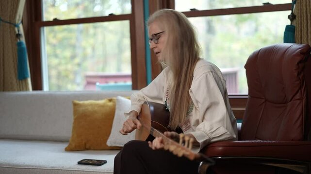 A woman with blonde hair and glasses sits in a brown leather chair, playing a saz instrument. She is wearing a white blouse and black pants. A white couch with yellow pillows is in the background.