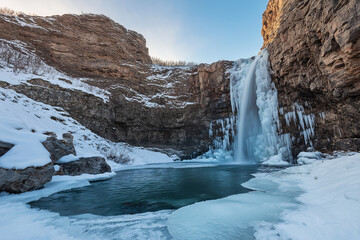 A photograph of a picturesque nature scene: an icy waterfall in Iceland, with snow and ice covering the ground, a blue sky, and the golden hour lighting