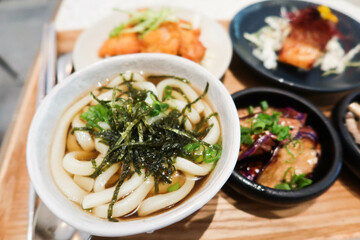 close up of soup udon with seaweed, mushroom appetiser, grilled salmon fish, eggplants and fried chicken in small plates bowls in a wooden tray