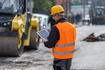 Construction worker in safety gear using a tablet to monitor progress at a busy urban site during...