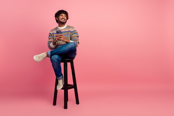 Full size portrait of nice young man hold phone look empty space sit stool isolated on pink color...