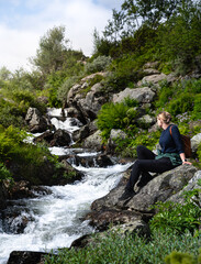 Woman enjoying a tranquil moment while watching the bubbling Silverfallet waterfall in a serene green setting, a female hiker sitting alone on a rock in the natural beauty of Dalarna, Sweden