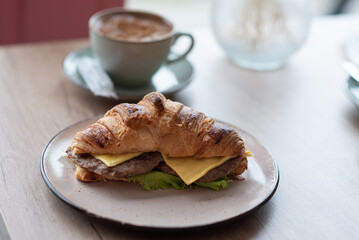 Close-up of a mug with coffee and a croissant with cheese and ham standing on a table in a cafe, near the window.