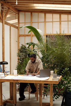Architect reviewing blueprints in lush, plant-filled workspace surrounded by greenery