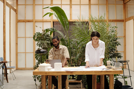 Two architects work on architectural designs at a wooden table surrounded by lush greenery