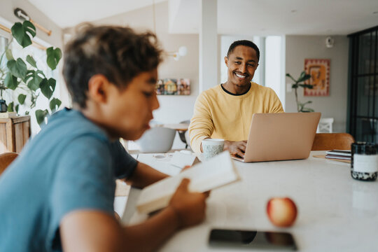 Happy father sitting with laptop while looking at son reading book at home