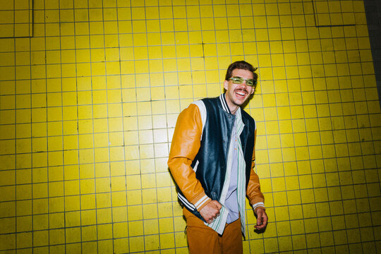 Portrait of happy young man in varsity jacket standing near yellow wall