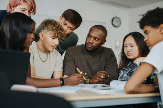 Teenage girl discussing with male teacher while sitting with classmates at desk