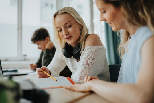 Smiling blond girl reading book while sitting by female teacher at desk in classroom