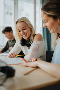 Happy blond girl reading book by female teacher sitting at desk