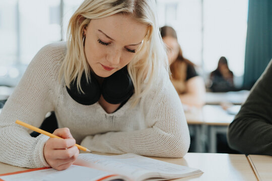 Blond teenage girl holding pencil while reading book at desk - Powered by Adobe