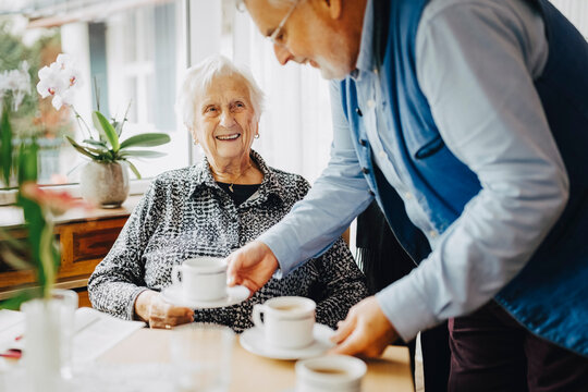 Elderly man serving tea to smiling senior man sitting at retirement home