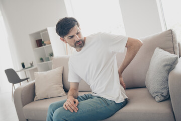 Young man experiencing back pain while sitting on a couch in a bright living room interior, highlighting discomfort and health awareness