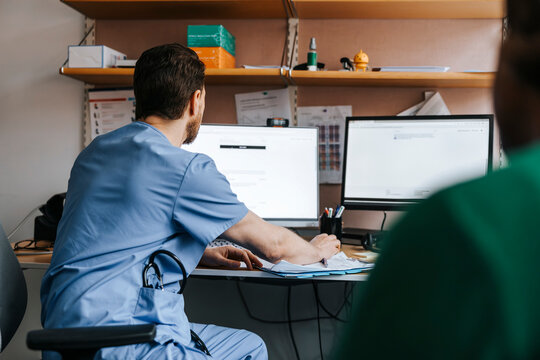 Male physician in blue scrubs checking medical records on computer while sitting in examination room at hospital